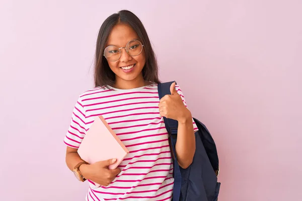 Chinese student woman wearing glasses backpack holding book over isolated pink background happy with big smile doing ok sign, thumb up with fingers, excellent sign