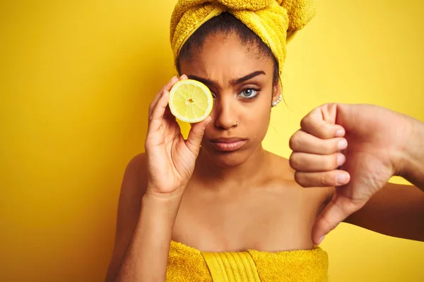 シャワーの後にタオルを着用したアフロ女性は 怒った顔で孤立した黄色の背景の上にスライスレモンを保持し 親指を下に嫌いを示す否定的なサイン 拒絶概念 — ストック写真