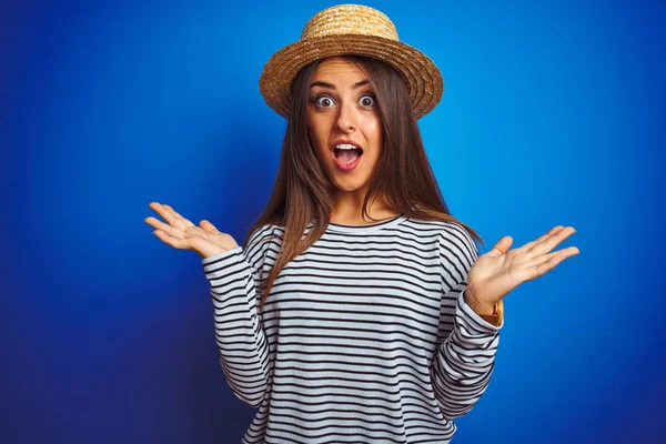 Young beautiful woman wearing navy striped t-shirt and hat over isolated blue background celebrating crazy and amazed for success with arms raised and open eyes screaming excited. Winner concept