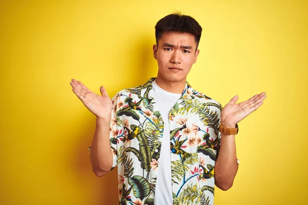 Asian chinese man on holiday wearing summer shirt over isolated yellow background clueless and confused expression with arms and hands raised. Doubt concept.