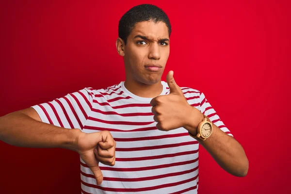Young handsome arab man wearing striped t-shirt over isolated red background Doing thumbs up and down, disagreement and agreement expression. Crazy conflict