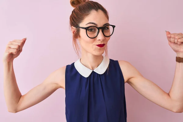 Redhead woman with pigtail wearing elegant dress and glasses over isolated pink background showing arms muscles smiling proud. Fitness concept.