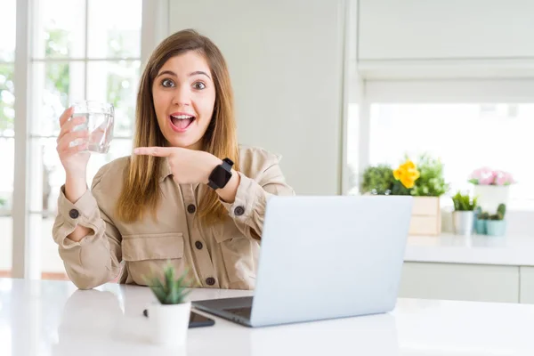 Beautiful young woman working with computer takes a break to drink glass of water very happy pointing with hand and finger