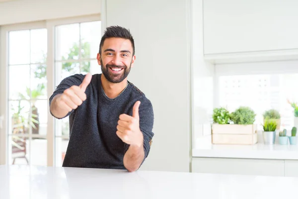 自宅でカジュアルなセーターを着ているハンサムなヒスパニックの男性は 手で肯定的なジェスチャーを行うことを承認し 親指は笑顔と成功のために幸せ カメラを見て 勝者のジェスチャー — ストック写真