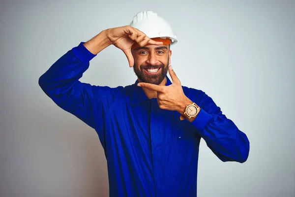 Handsome indian worker man wearing uniform and helmet over isolated white background smiling making frame with hands and fingers with happy face. Creativity and photography concept.