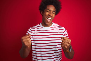 Afro saçlı genç afro-amerikan adam izole kırmızı arka plan üzerinde çizgili t-shirt giyen çok mutlu ve heyecanlı kolları kaldırdı ile kazanan jest yapıyor, gülümseyerek ve başarı için çığlık. Kutlama konsepti.