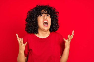 Kıvırcık saçlı genç arap kadın izole kırmızı arka plan üzerinde rahat t-shirt giyen deli ve deli bağırarak ve agresif ifade ve kollar kaldırdı ile bağırıyor. Hayal kırıklığı kavramı.