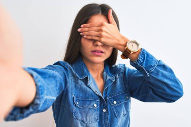 Denim gömlek giyen güzel kadın, gözleri eliyle kaplayan, ciddi ve hüzünlü görünen izole beyaz arka plan üzerinde kamera yla selfie çekiyor. Görme, gizleme ve reddetme kavramı