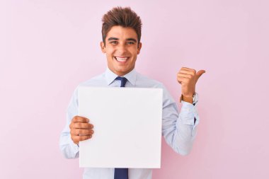 Genç yakışıklı işadamı izole pembe arka plan üzerinde duran afiş tutan işaret ve mutlu yüzü gülümseyerek yan başparmak ile gösteren