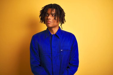 Izole sarı arka plan üzerinde mekanik üniforma giyen dreadlocks ile Afro işçi adam yan bakarak gülümseyerek ve düşünme uzak bakıyor.