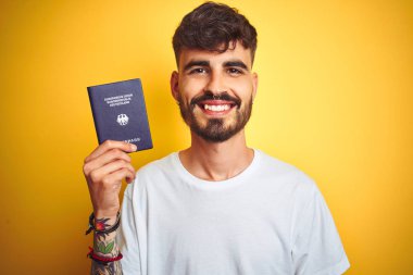 Izole sarı arka plan üzerinde Alman Almanya pasaportu giyen dövmeli genç adam ayakta ve dişleri gösteren kendinden emin bir gülümseme ile gülümseyen mutlu bir yüz ile