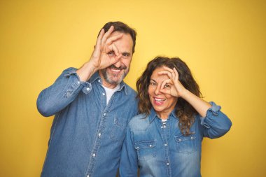Güzel orta yaş çift birlikte izole sarı arka plan üzerinde duran el gülümseyerek ok jest yapıyor, mutlu yüz ile parmaklardan bakan göz.