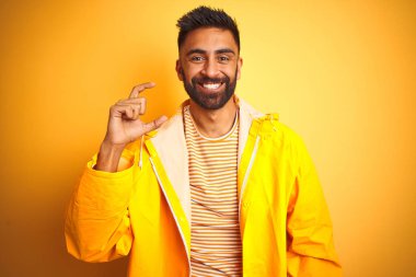 Genç hintli adam izole sarı arka plan üzerinde duran yağmurluk giyen gülümseyerek ve emin el parmakları ve kamera görünümlü küçük boyutlu işareti yapıyor gesturing. Ölçü kavramı.