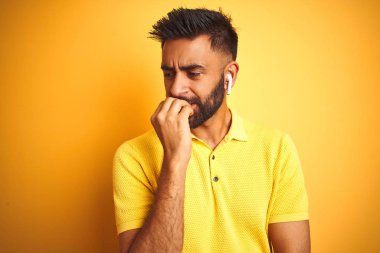 Genç Hintli adam izole sarı arka plan üzerinde duran kulaklık kullanarak müzik dinlerken stresli ve ağız tırnakısırma elleri ile sinirli görünümlü. Anksiyete sorunu.