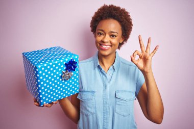 Pembe izole arka plan üzerinde doğum günü hediyesi tutan Afro-amerikan kadın parmakları ile ok işareti yapıyor, mükemmel sembolü
