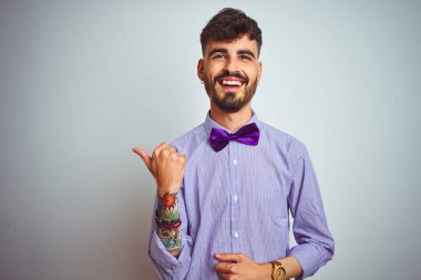 Dövme mor gömlek ve izole beyaz arka plan üzerinde papyon giyen genç adam mutlu yüz görünümlü gülümseyerek ve başparmak ile yan işaret.