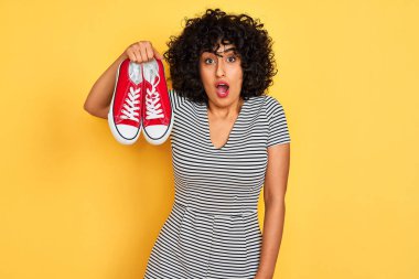 Kıvırcık saçlı genç arap kadın izole sarı arka plan üzerinde rahat spor ayakkabı tutan sürpriz bir yüz ile şok korkmuş, korku ve korku ifade ile heyecanlı