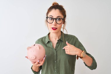 Kendine parmak işaret sürpriz yüzü ile izole beyaz arka plan üzerinde kumbara tutan gözlük takan güzel kızıl saçlı kadın