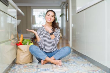 Genç kadın bir kağıt çanta ile mutfak katında oturan taze bakkaliye hayran dolu ve kamera için gülümseyen el ile sunmak ve parmak ile işaret.