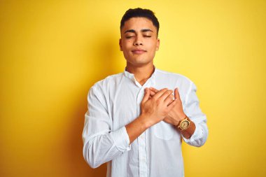 İzole sarı arka plan üzerinde duran zarif gömlek giyen genç Brezilyalı işadamı kapalı gözleri ve yüzünde minnettar jest ile göğüs üzerinde gülümseyerek. Sağlık kavramı.