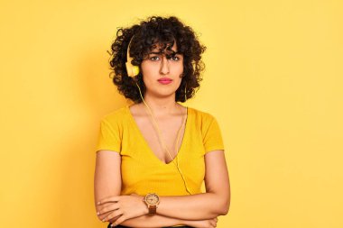 Kıvırcık saçlı Arap kadın izole sarı arka plan şüpheci ve sinir üzerinde kulaklık kullanarak müzik dinlerken, çapraz kolları ile yüzünde ifade onaylamayan. Negatif kişi.