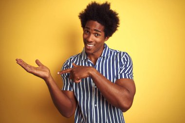 Afro saçlı, izole edilmiş sarı arka planın üzerinde duran çizgili gömlek giyen Amerikalı adam, el ile sunarken ve parmakla işaret ederken kameraya gülümsüyor ve gülümsedi.