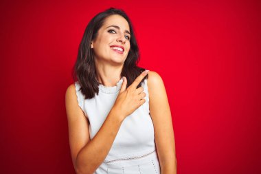 Kırmızı izole arka plan üzerinde duran beyaz elbise giyen genç güzel kadın yüzünde mutlu ve doğal ifade ile yan yana el ve parmak ile işaret eden yüz bir gülümseme ile neşeli