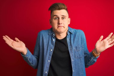 Denim gömlek giyen genç yakışıklı adam izole kırmızı arka plan üzerinde duran habersiz ve kolları ve elleri kaldırdı ile karışık ifade. Şüphe kavramı.