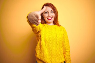 Güzel kızıl saçlı kadın izole sarı arka plan üzerinde duran kış kazak giyen tebrik ve karşılama olarak dostça sunan samimi gülümseyerek. Başarılı iş.
