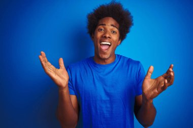 Afro saçlı afro saçlı adam izole mavi arka plan üzerinde duran t-shirt giyen deli kutluyor ve kollarını kaldırdı ve açık gözleri heyecan çığlık ile başarı için hayran. Kazanan kavramı