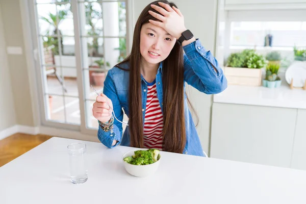 Beautiful Asian woman eating green fresh broccoli stressed with hand on head, shocked with shame and surprise face, angry and frustrated. Fear and upset for mistake.