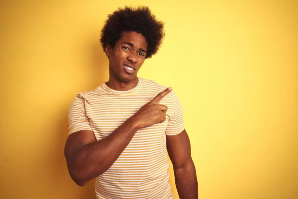 American man with afro hair wearing striped t-shirt standing over isolated yellow background Pointing aside worried and nervous with forefinger, concerned and surprised expression