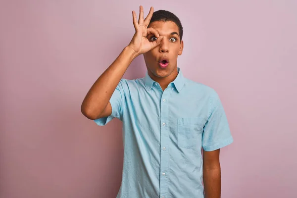 孤立したピンクの背景の上に立つ青いシャツを着た若いハンサムなアラブの男は 驚いた顔 指を通して目を見てショックを受けたOkジェスチャーをやっています 信じられない表現 — ストック写真