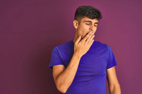 Young indian man wearing t-shirt standing over isolated purple background bored yawning tired covering mouth with hand. Restless and sleepiness.