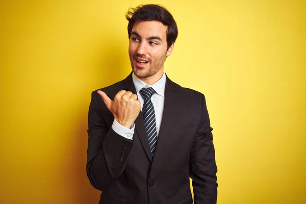 スーツとネクタイを着た若いハンサムなビジネスマンは 親指を上げて横を向いて幸せそうな顔で微笑む孤立した黄色の背景の上に立っている — ストック写真