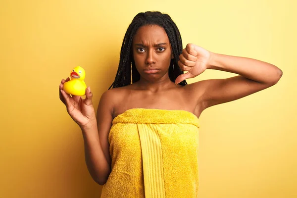 African american woman wearing shower towel holding toy duck over isolated yellow background with angry face, negative sign showing dislike with thumbs down, rejection concept