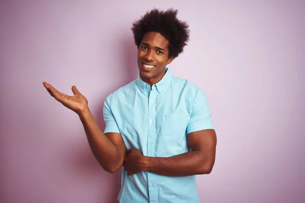 青いシャツを着たアフロヘアの若いアメリカ人男性が カメラを見て手のひらを指差して陽気な笑顔を浮かべて 孤立したピンクの背景の上に立っている — ストック写真