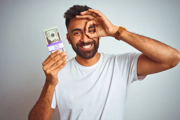 年轻的印度男子拿着美元站在孤立的白色背景与快乐的脸微笑做Ok标志与手在眼睛上通过手指看 — 图库照片