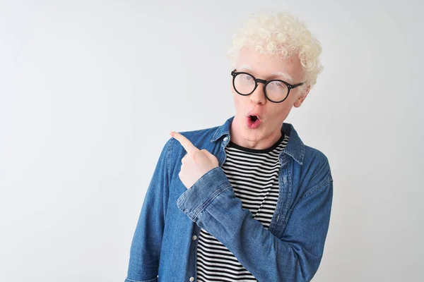Young albino blond man wearing denim shirt and glasses over isolated white background Surprised pointing with finger to the side, open mouth amazed expression.