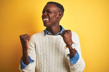 Denim gömlek ve beyaz kazak giyen Afro-amerikan adam izole sarı arka plan üzerinde kutlayan şaşırmış ve kollarını kaldırdı ve gözleri kapalı başarı için şaşırttı. Kazanan kavramı.