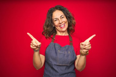 Orta yaş yaşlı kadın kırmızı izole arka plan üzerinde önlük üniforma giyen farklı yönlere parmakları ile işaret emin gülümseyerek. Reklam için kopya alanı