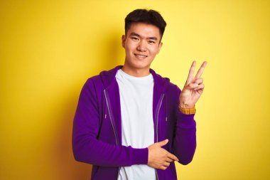 Mor sweatshirt giyen genç Asyalı çinli adam izole sarı arka plan üzerinde duran mutlu yüz ile gülümseyerek zafer işareti yapıyor kameraya göz kırparak. İki numara.