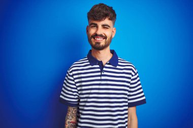 Seksi ifade, neşeli ve mutlu yüz ile kameraya bakarak izole mavi arka plan üzerinde duran çizgili polo giyen dövme giyen genç adam.