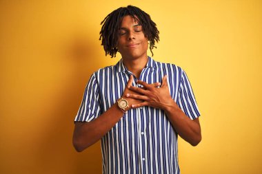 Dreadlocks ile rahat çizgili t-shirt izole sarı arka plan üzerinde kapalı gözleri ve yüzünde minnettar jest ile göğüs elleri ile gülümseyen giyen Afro adam. Sağlık kavramı.