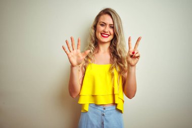 Beyaz izole arka plan üzerinde duran sarı t-shirt giyen genç güzel kadın gösteren ve emin ve mutlu gülümserken parmakları ile yedi numara işaret.