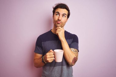 Izole pembe arka plan ciddi yüz soru hakkında düşünme dövme içme fincan ile genç yakışıklı adam, çok karışık bir fikir