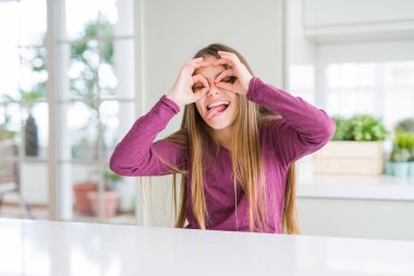 Beyaz masadaki güzel genç kız çocuk dürbün gibi tamam jest yapıyor dilini dışarı sokuyor, gözleri parmaklardan bakıyor. Çılgın ifade.
