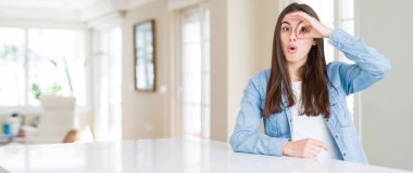 Güzel genç kadının evde beyaz masada oturan geniş açı resim sürpriz yüz ile şok ok jest yapıyor, göz parmaklar bakarak. İnanılmaz ifade.