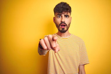 Çizgili t-shirt giyen dövme giyen genç adam izole sarı arka plan üzerinde duran hoşnut olmayan ve kameraya sinirli işaret, kızgın ve öfkeli size