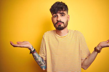 Çizgili t-shirt giyen dövme giyen genç adam izole sarı arka plan üzerinde duran habersiz ve kolları ve elleri ile karışık ifade kaldırdı. Şüphe kavramı.